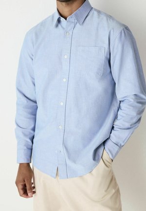 Homme portant une chemise bleu clair à boutons avec manches longues et un pantalon beige, debout avec une main dans la poche.