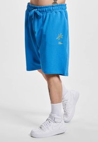 Shorts din bumbac albaștri cu un elastic la talie, având brodat o palmier în verde. Purtați cu adidași albi cu gânduri înalte.