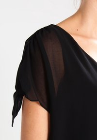 Blusa negra con mangas abullonadas transparentes y escote en forma de V. La tela parece ligera y suave, con un diseño suelto y fluido.