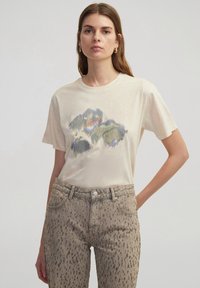 Camiseta de manga corta beige claro con un colorido diseño gráfico abstracto. Combinada con jeans beige texturizados con patrones moteados oscuros.