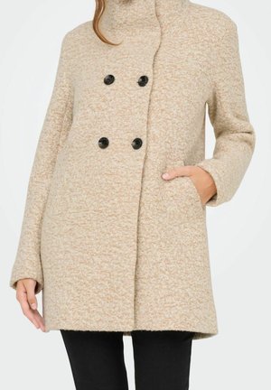 Femme portant un manteau croisé en laine beige avec des boutons noirs, une main dans la poche, associée à un pantalon noir.