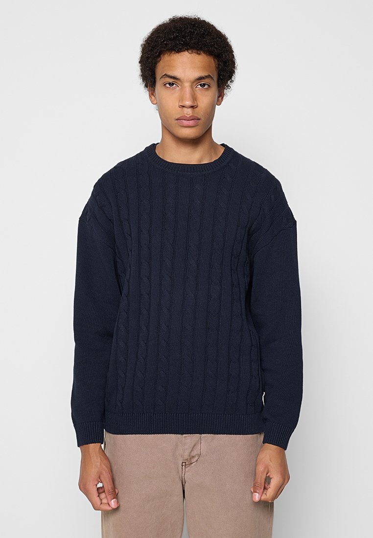 Junger Mann mit lockigem Haar, der einen navyblauen Pullover mit Kabelmuster und beigen Hosen trägt, steht vor einem einfarbigen hellgrauen Hintergrund.