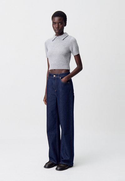Suéter gris corto con un diseño de cuello de perlas, combinado con jeans de pierna ancha en azul oscuro. El modelo lleva zapatos negros y está de pie contra un fondo claro.