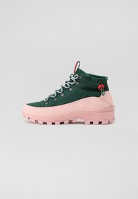 Bota de senderismo verde y rosa con parte superior texturizada, cordones grises y una suela rosa distintiva. Presenta un acento rojo y ojales hexagonales.