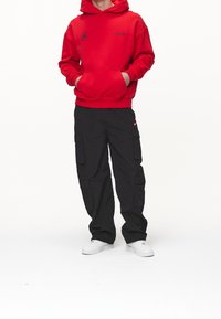 Röd huvtröja med framficka och svart logotyp, i kombination med svarta cargobyxor med sidofickor och vita sneakers.