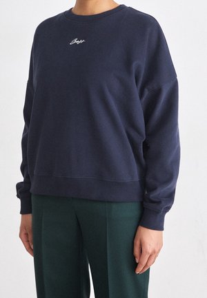 Persoon draagt een losse marineblauwe trui met een klein wit scriptlogo op de borst en donkergroene broek, staand tegen een effen achtergrond.