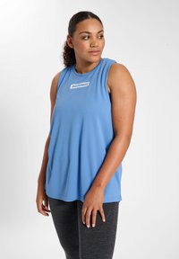 Sleeveless blue athletic tank top in soft fabric with a rounded neckline, featuring a white logo in the center. Paired with textured black leggings.