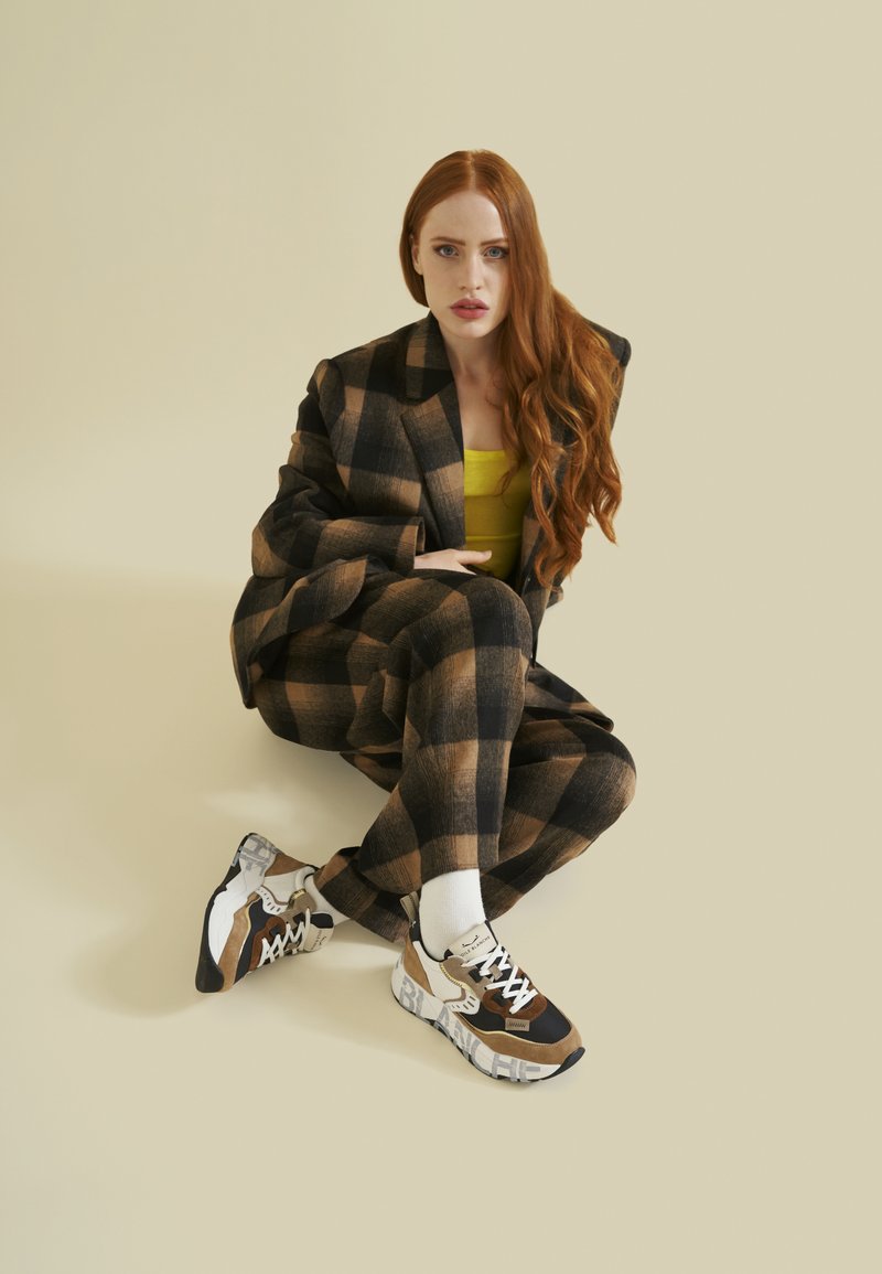 Completo a quadri marrone e nero con giacca aderente e pantaloni rilassati, abbinato a sneakers multicolori e calze bianche. Parte superiore gialla visibile.