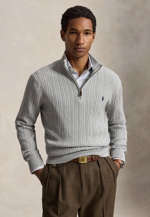 Homme portant un pull tricoté gris clair avec fermeture éclair quart de zip sur une chemise rayée et un pantalon marron à carreaux, les mains dans les poches, sur fond neutre.