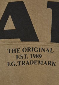 Tessuto beige con testo nero audace. Il testo include "L'ORIGINALE," "FONDATO NEL 1989," e "EG. MARCHIO REGISTRATO." La texture appare liscia e tessuta.