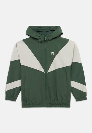 Chaqueta verde con patrones triangulares de color gris claro, cremallera frontal, capucha y puños elásticos. Cuenta con bolsillos laterales y un logotipo en el pecho.