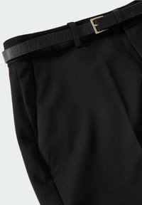 Pantalon noir sur mesure avec plis devant et une fine ceinture noire texturée avec une boucle dorée sur fond clair.