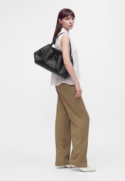 Femme en blouse blanche sans manches et pantalon marron à jambes larges portant un grand sac à main en cuir noir sur l'épaule, sur un fond uni.