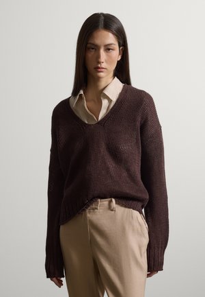 Donna con capelli lisci e scuri indossa un maglione oversize marrone scuro sopra una camicia beige a righe con colletto e pantaloni beige a vita alta.
