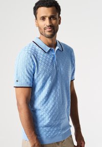 Světle modrá pletená polo košile s texturovaným čtvercovým vzorem, s kontrastním tmavým límcem a manžetami, předními knoflíky a žebrovaným lemem.