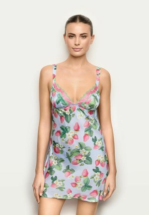 Mujer con un vestido tipo slip azul claro con patrones de fresas rosas y hojas verdes, posando contra un fondo liso y claro.