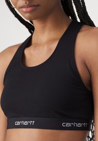 Black sports bra made of soft fabric, featuring a rounded neckline and a wide band with white "Carhartt" logo. Smooth texture, minimal design.