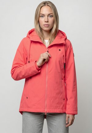 Frau mit langen blonden Haaren, die eine korallrosa Kapuzenjacke trägt und diese vor einem schlichten, hellen Hintergrund zumacht.