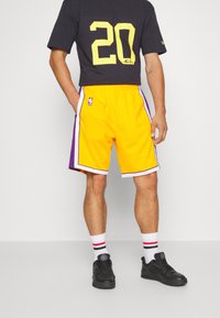 Pantaloni da basket gialli con accenti viola, strisce laterali e logo NBA. Abbinati a una maglietta nera con un grande numero 20 giallo.