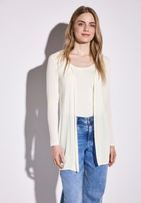 Cardigan à manches longues, ouvert à l'avant, en tissu crème clair, superposé sur un haut blanc, associé à un jean bleu à jambes larges. Texture lisse, design minimaliste.