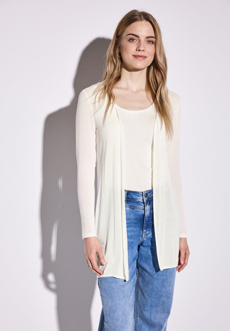 Cardigan à manches longues, ouvert à l'avant, en tissu crème clair, superposé sur un haut blanc, associé à un jean bleu à jambes larges. Texture lisse, design minimaliste.