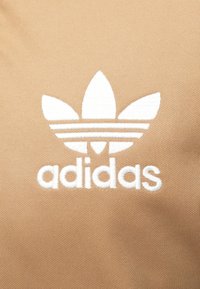 Μπεζ ύφασμα με λευκό κεντημένο λογότυπο Adidas και τρία φύλλα περιμετρικά, με το όνομα της μάρκας παρακάτω. Λείο, πυκνό ύφασμα.