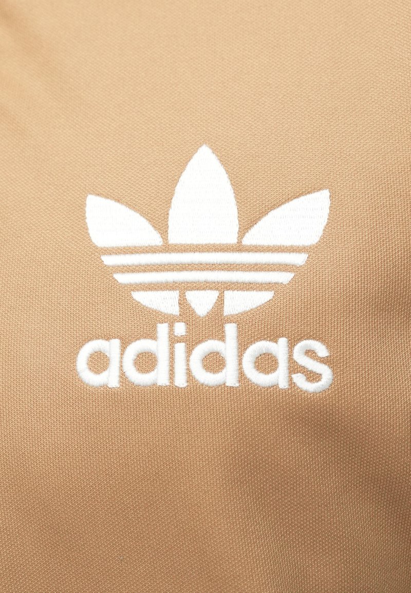 Beige Textil mit einem weißen gestickten Adidas-Logo, das drei blattähnliche Akzente und den Markennamen darunter zeigt. Glattes, eng gestricktes Material.