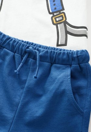 Baumwoll-T-Shirt mit weißem Grund und grafischen blauen Trägern, kombiniert mit blauen Shorts mit elastischem Bund und Seitentaschen.