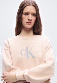 Νεαρή γυναίκα με μακριά καστανά μαλλιά, φορώντας ένα ανοιχτό ροζ φούτερ Calvin Klein, στέκεται με τα χέρια σταυρωμένα μπροστά σε ένα απλό φόντο.