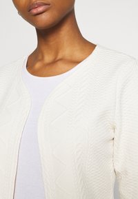 Kartenigan in Cremefarbe mit einem strukturierten Zickzackmuster, offenem Frontdesign und rundem Ausschnitt, getragen über einem weißen Oberteil.