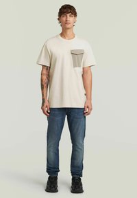 T-shirt in cotone beige con tasca grigia, maniche corte e scollatura rotonda, abbinata a jeans blu e sneakers nere.