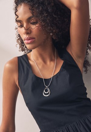 Woman with curly hair wearing a sleeveless black top and a silver necklace with overlapping oval pendants, arm raised behind her head.
