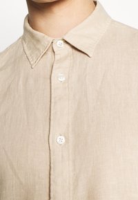 Camicia di lino beige con colletto classico, quattro bottoni bianchi davanti e un tessuto testurizzato con una finitura naturale.