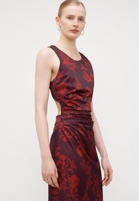 Rochie bordeaux fără mâneci, cu un model floral în nuanțe de roșu, având decupaje laterale și material drapat la talie pentru un plus de textură și design.