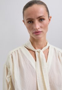 Blusa color crema confeccionada en una tela ligera, con un lazo en el cuello, mangas plegadas y una silueta relajada. Textura suave y caída agradable.