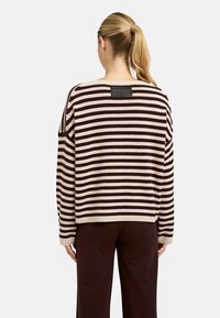 Frau mit blonden Pferdeschwanz, die einen braun-beige gestreiften Pullover und dunkle braune Hosen trägt, mit dem Rücken zu einem weißen Hintergrund.