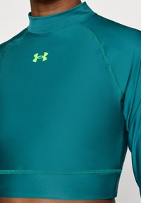 Αθλητική μπλούζα με μακριά μανίκια σε τυρ κουάζ χρώμα, κατασκευασμένη από λείο ύφασμα. Διαθέτει λαιμόκοψη mock neck, εφαρμοστό σχέδιο και ένα μικρό κίτρινο λογότυπο Under Armour στο στήθος.