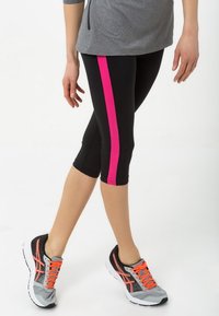 Czarne legginsy capri z różowym paskiem po boku, zestawione z szaro-czarnymi butami sportowymi z pomarańczowymi akcentami. Miękki, elastyczny materiał.