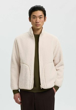 Homme portant une veste polaire crème zippée sur un pull vert, avec un pantalon foncé, debout devant un fond clair uni.