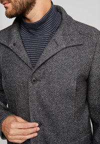 Abrigo gris texturizado con un gran cuello, con cierre de botón oscuro. Un cuello alto a rayas azules marinas asoma por debajo.