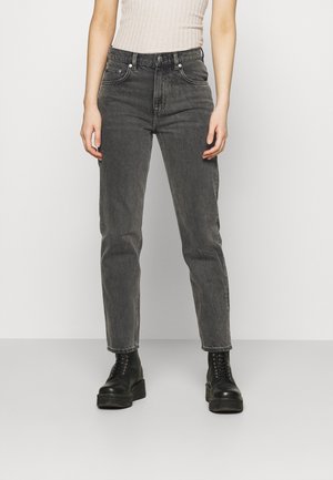 Kvinde iført højtaljede, afblegede sorte jeans og sorte snørestøvler med plateau, stående mod en ensartet lys baggrund.