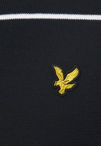 Tissu noir tricoté avec une bande blanche et un logo aigle brodé en or à l'avant. Le design est simple et épuré.