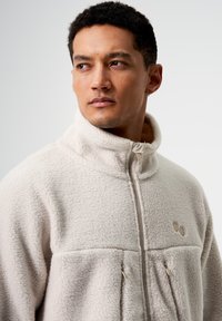 Cremefarbene Fleecejacke mit hohem Kragen, Reißverschluss, zwei Fronttaschen und dezentem Branding. Weiche Textur und lockere Passform.