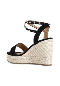 Sandalo con zeppa in suede nero e piattaforma in juta intrecciata. Presenta un design con cinturini e borchie in tonalità oro, e cinturini alla caviglia regolabili.