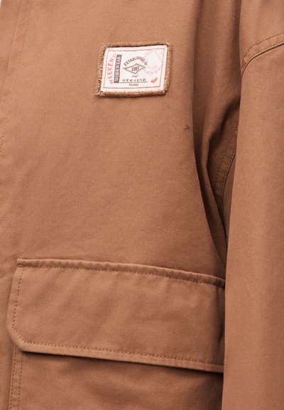 Καφέ μπουφάν με μπροστινή τσέπη με καπάκι και ορθογώνια λευκή ετικέτα με την επιγραφή "Weekend Workwear Established 1983 Italy Weekend".