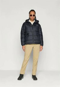 Chaqueta acolchada negra con capucha, con cremallera en el frontal, diseño acolchado y un pequeño logo en el pecho. Combinada con pantalones caqui y botas negras.