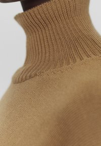 Zbliżenie na jasnobrązowy sweter z golfem, ukazujące teksturę dzianiny oraz detale kołnierza wokół szyi i ramion.