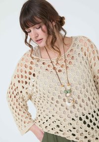 Pull en crochet beige avec de grandes ouvertures circulaires, manches trois-quarts, associé à un collier multi-couches ornée de perles en bois et en marbre.
