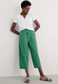 Fehér V-kivágású póló, zöld, rövidített, gumis derekú nadrággal párosítva. Világosszürke szandálokkal viseli, keresztben futó pántokkal és texturált talppal.