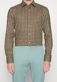 Ein Hemd mit Knopfleiste im braun-schwarzen Hahnentrittmuster, mit klassischem Kragen und langen Ärmeln. Kombiniert mit hellblauen Hosen.